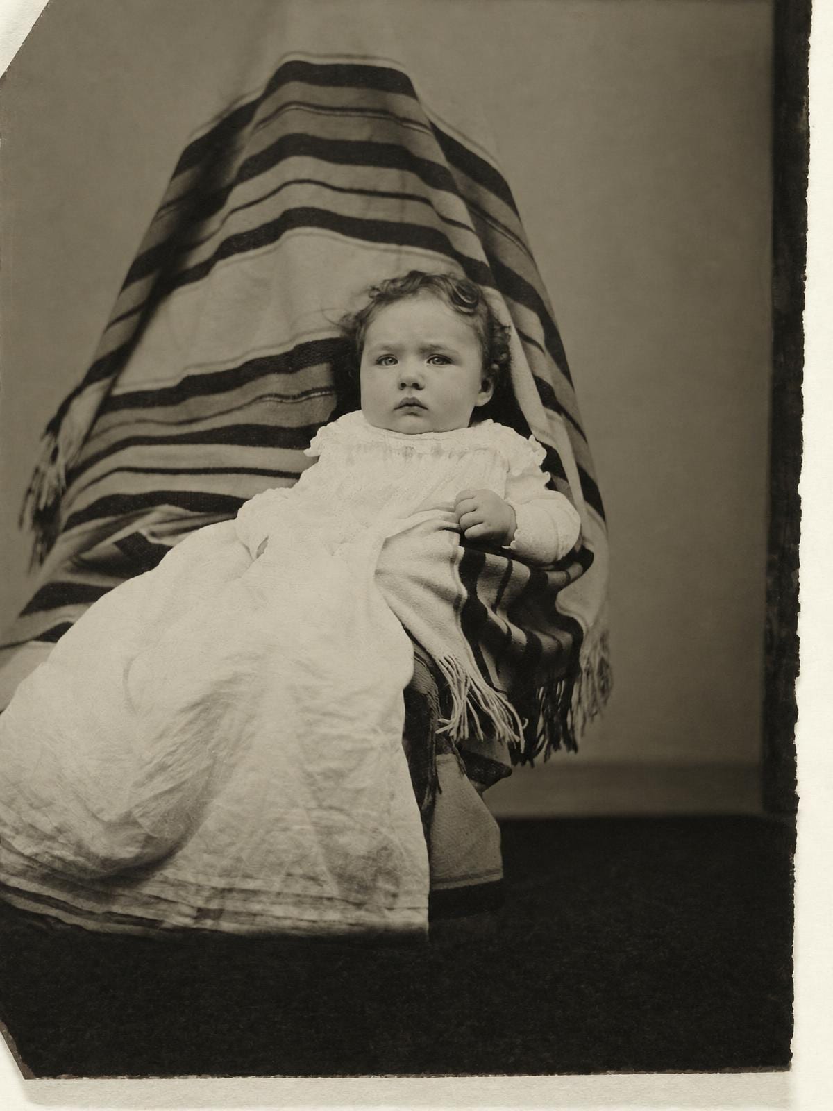 A tintype of a little girl in a long white gown on the lap of a figure hidden under a striped shawl, scratched, tarnished and spotted with rust at the right edge, beside the same photo repaired with the shawl still in place