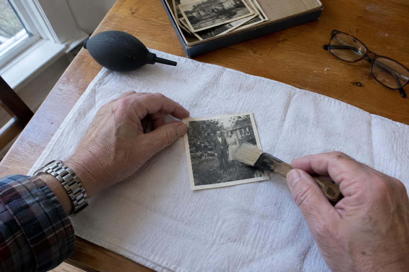 Hands holding a small black-and-white snapshot flat on a white towel by its border and sweeping a wide soft brush across it, with a rubber bulb blower beside the towel
