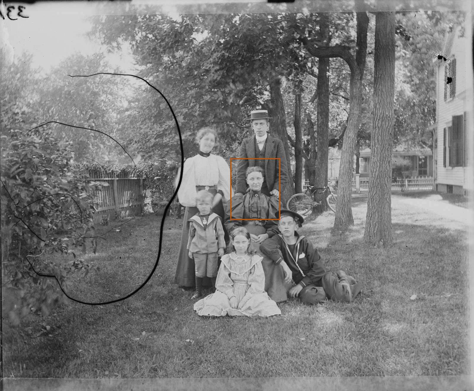 An 1898 black-and-white photo of a family of six on a lawn under trees, with an orange frame around the head and shoulders of the woman seated in the middle.
