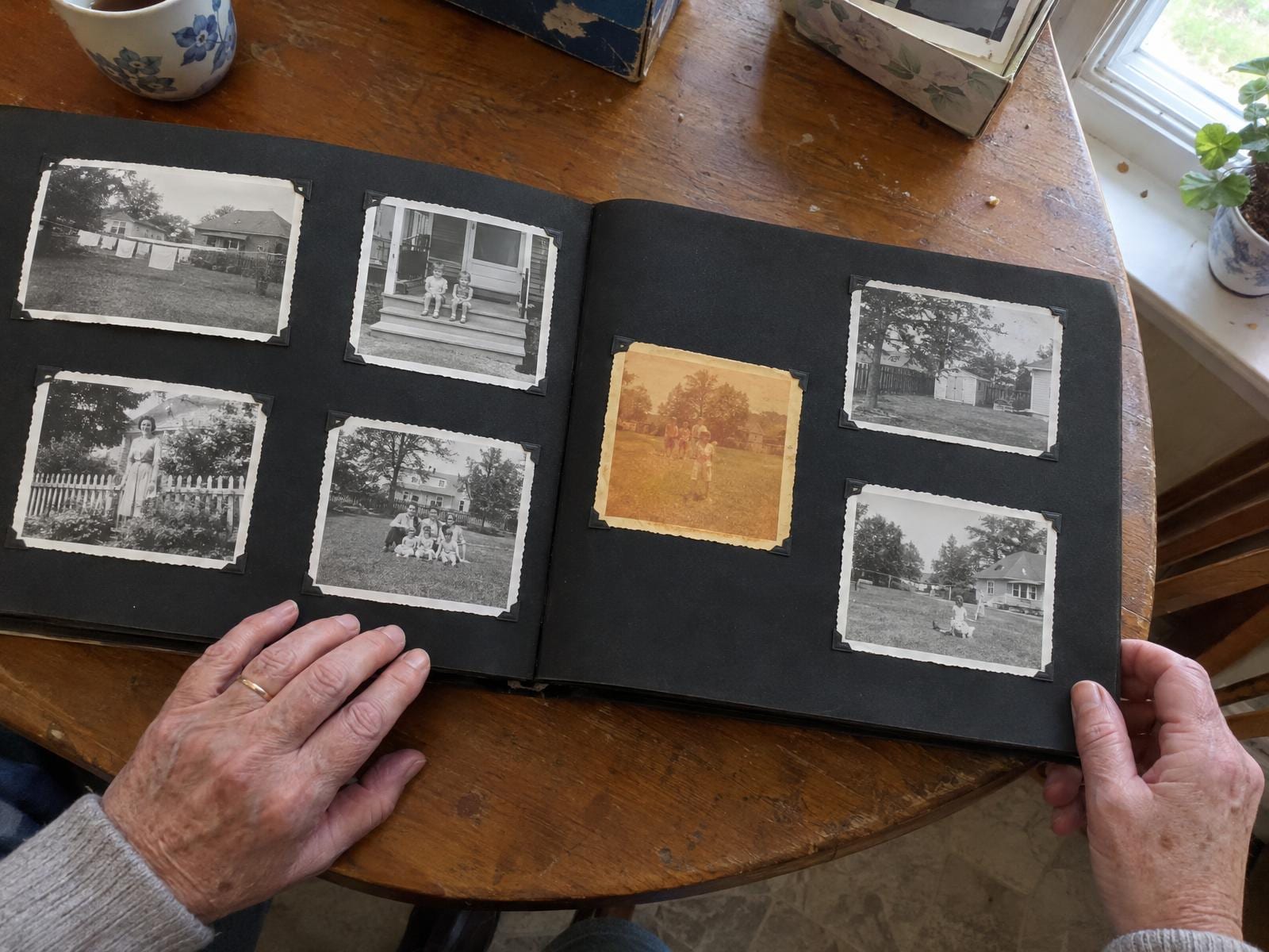 An open family album on a kitchen table: black pages of small black-and-white snapshots held by photo corners, and one early color print faded to an orange-yellow cast, with an older woman's hands holding the pages flat