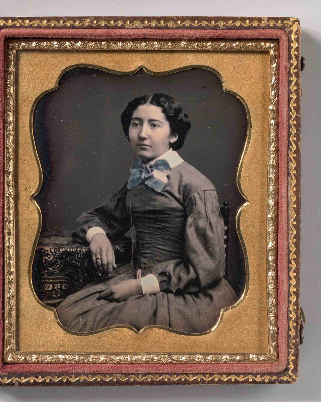 An 1850s daguerreotype in its gilt case: a seated young woman in a gray dress, with her bow tinted blue, her cheeks pink and her ring touched with gold by hand