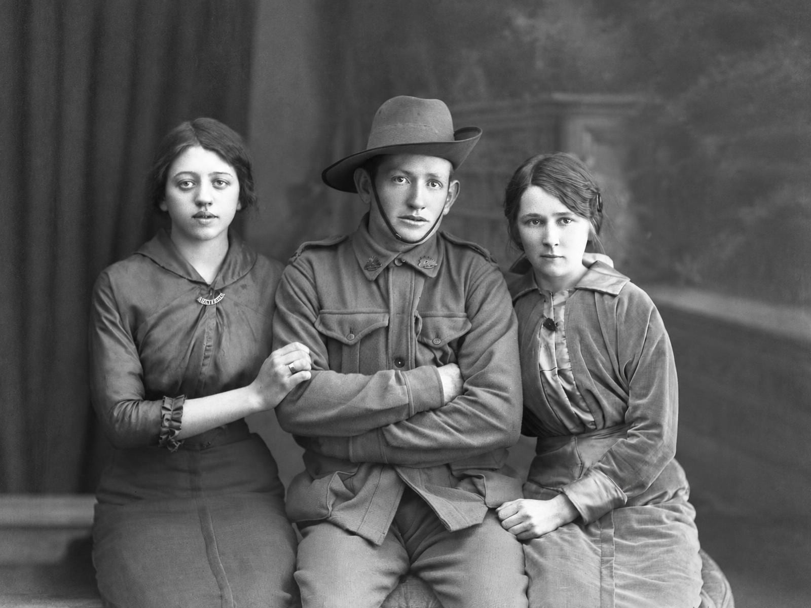 A 1910s studio portrait of a soldier seated between two young women, with water damage eating into the edges, before and after a restore that kept it black and white
