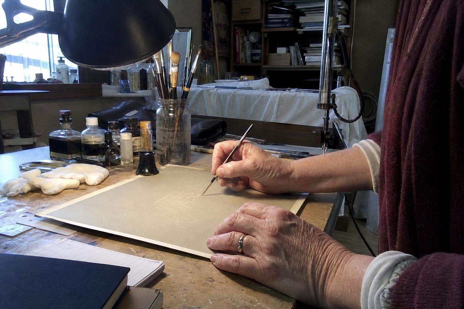 A retoucher's hands working on a print with a fine brush under a desk lamp