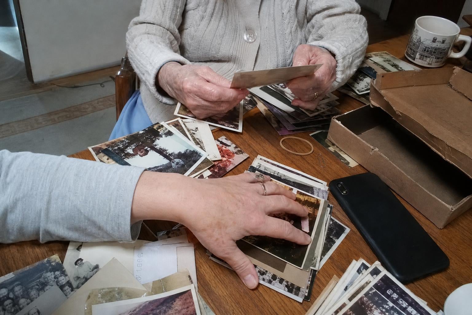 Hands sorting a shoebox of old family photos on a table beside a phone